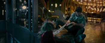 Brandon Sklenar tente de tirer à lui une nappe sur laquelle se tient une femme blonde, suspendue dans le vide ; scène de nuit, au bord d'un salon dont la large vitre a été brisée, dans le film Drop game.