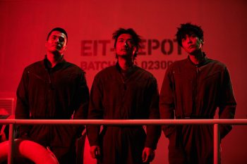 Les trois adolescents du film Escape From the 21st Century en tenue d'usine, sur une plateforme d’entrepôt plongé dans une lumière rouge ; ils regardent le plafond, la mine flottante.