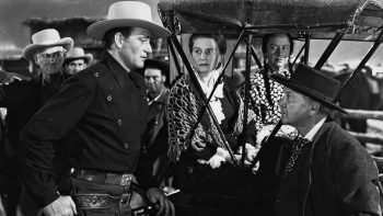 John Wayne debout face à un grand propriétaire terrien de Fargo, sous le regard d'hommes de mains prêts à agir ; scène du film La femme du pionnier.