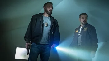 Matt Damon et Ben Affleck, une torche à la main, enquêtent et scrutent dans une pièce aux murs blancs ; scène du film The rip.