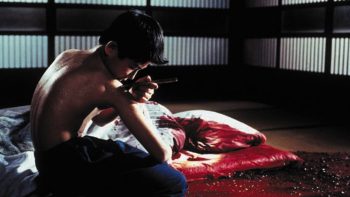 Un jeune homme, dans une chambre japonaise, à côté d'un drap blanc maculé de sang, se fait lui-même un tatouage sur l'épaule ; scène de Graine de yakuza.