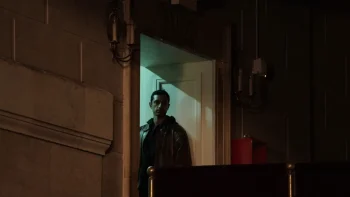 Riz Ahmed au premier étage d'un théâtre, sur le palier d'une porte de service, observant la scène de loin dans le film L'intermédiaire.