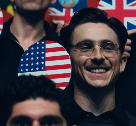 Timothée Chalamet souriant en portant une raquette de ping pong peinturé du drapeau des États-Unis dans Marty Supreme.