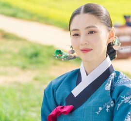 Han So-hee en tenue traditionnelle, souriante, sur un fond de petit chemin de campagne lumineux.