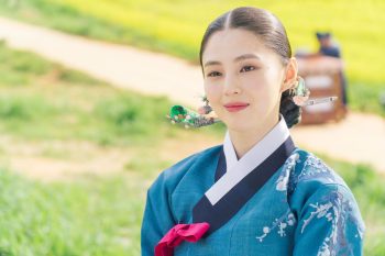 Han So-hee en tenue traditionnelle, souriante, sur un fond de petit chemin de campagne lumineux.