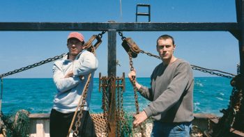 Deux hommes sont sur le pont d'un bateau de pêche ; celui de gauche porte une casquette rouge et sweat blanc ; celui de droite a les cheveux coupés très courts et porte un pull gris ; tous deux s'appuient ou tirent sur des chaînes rouillées ; on aperçoit la mer bleue en arrière-plan, de même qu'un ciel sans nuages ; plan du film ROSE OF NEVADA, projeté à l'Offscreen Film Festival 2026.
