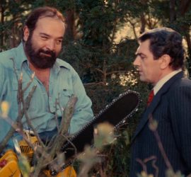 Un homme montre sa tronçonneuse à un autre en souriant dans ce qui semble être un jardin ; plan du film Le sadique à la tronçonneuse.