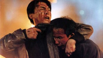 Tony Leung, le visage blessé, tient sous son bras son ami en poitant son revolver contre son crâne ; derrière eux un imposant feu ; scène finale du film Une balle dans la tête.