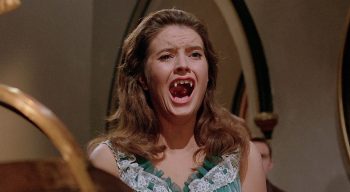 Une femme aux longs cheveux blonds et portant une robe bleue découvre ses dents de vampires devant un miroir ; plan du film LE BAISER DU VAMPIRE.