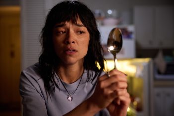 Une jeune femme au regard apeuré observe le dos d'une cuillère qu'elle tient dans ses mains ; elle porte des cheveux noirs avec une frange ; elle est vêtue d'un haut bleu et un collier pend autour de son cou ; on devine qu'elle doit se trouver dans une cuisine peu éclairée ; en arrière-plan on image un frigo à la porte ouverte ; plan du film SACCHARINE, projeté au BIFFF 2026. 
