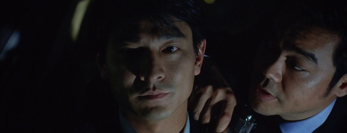 un homme avec un revolver dans la main parle à un autre homme qui regarde la camera, l'image est sombre ; plan issu de Running out of time.