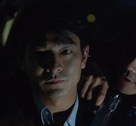 un homme avec un revolver dans la main parle à un autre homme qui regarde la camera, l'image est sombre ; plan issu de Running out of time.