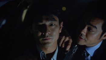 un homme avec un revolver dans la main parle à un autre homme qui regarde la camera, l'image est sombre ; plan issu de Running out of time.