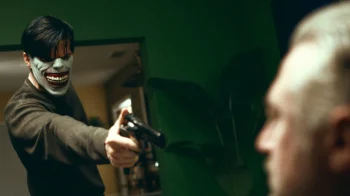 Un homme aux cheveux courts et portant un masque de clown braque un revolver en direction d'un autre homme ; l'autre homme est en amorce du plan et on imagine qu'il est d'un certain âge ; on n'aperçoit qu'une partie du visage flou de l'homme pris en joue ; l'homme ciblé a des cheveux blancs ; la pièce est sombre ; plan du film APPOFENIACS, projeté au BIFFF 2026.