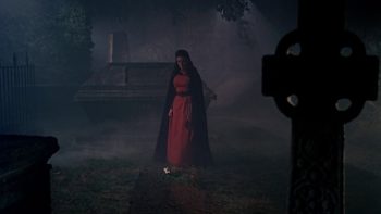 Dans un cimetière de nuit, une femme en robe rouge et portant une cape sombre déambule entre des tombes ; le cimetière est plongé dans une brume épaisse ; plan du film LE BAISER DU VAMPIRE.