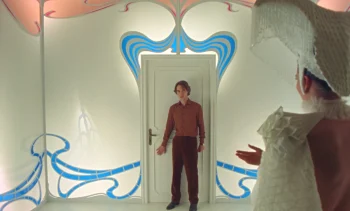 Un homme se tient devant une porte blanche ; la pièce semble sortie d'un rêve ; les murs sont blancs et parcourus de courbes bleues ; l'ambiance qui se dégage est particulièrement onirique ; en amorce du plan, on aperçoit le dos d'une dame vêtue de vêtements blancs ; plan du film ORFEO, projeté au BIFFF 2026.