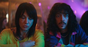 Une femme et un homme sont côte à côte dans un bar dansant ; ils apparaissent en plan rapproché et occupent tout le cadre ; la femme (Asia Argento) a les cheveux noirs et porte un haut jaune ; elle regarde vers le bas, les bras croisés ; l'homme a de longs cheveux bouclés et une moustache ; il a les yeux ébahis et semble surpris ; plan du film PLUS FORT QUE LE DIABLE de Graham Guit.