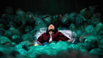 Une femme est allongée au milieu de sacs de couleur turquoise ; on imagine que ces sacs sont des sacs de déchets ; elle a les yeux ouverts et semble pensive ; elle porte une veste sombre au-dessus d'un vêtement clair ; la femme en question semble d'origine asiatique ; plan du film SACCHARINE, projeté au BIFFF 2026.