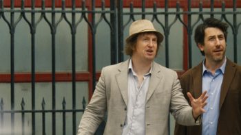 Deux hommes se tiennent à gauche de l'image ; le premier porte un chapeau et une veste beiges ; le deuxième porte un veston au-dessus d'une chemise bleu ciel ; derrière eux, on devine une grille en fer forgé ; plan du film NIRVANNA THE BAND THE SHOW THE MOVIE, projeté au BIFFF 2026.