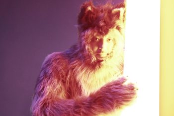 Un homme déguisé en chien se tient en plein milieu de l'image ; il porte un costume composé de longs poils ; son visage apparaît, mais le costume de chien lui recouvre l'ensemble du corps ; le mur derrière lui est de couleur mauve ; son visage est éclairé par une barre de lumière située sur la droite de l'image ; plan du film GOSH!!, projeté au BIFFF 2026.