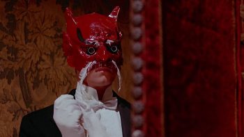 Un personnage en costume porte un étrange masque de couleur rouge ; il porte des gants blancs, une chemise blanche, un nœud papillon blanc et une veste noire ; on n'aperçoit que son menton, le masque rouge lui voilant le reste du visage ; plan du film LE BAISER DU VAMPIRE.