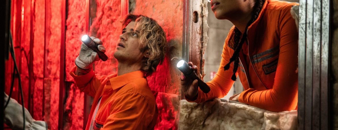 Un homme incarné par Joe Keery et une femme en tenues orange tenant des lampes torches dans un endroit désaffecté.