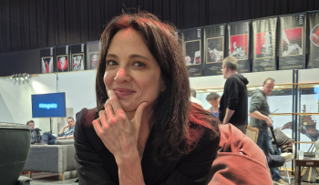 Asia Argento se tient le menton et regarde l'appareil photo ; elle est assise sur une chaise ou un fauteuil et, derrière elle, apparaissent des festivaliers ainsi qu'une série d'affiches fixées en hauteur ; photo prise au BIFFF 2026.