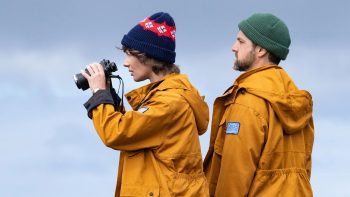 Un homme et une femme apparaissent en avant-plan tandis qu'on devienne la mer derrière eux ; tous deux portent un bonnet ainsi qu'une veste jaune, capuchon rabattu ; la femme tient des jumelles entre les mains, qu'elle semble porter vers ses yeux ; les deux personnages regardent vers la gauche de l'image ; plan du film YESTERDAY ISLAND, projeté au BIFFF 2026.