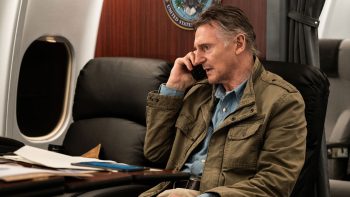 Liam Neeson au téléphone assis dans un avion avec plein de papiers posés sur une tablette devant lui.