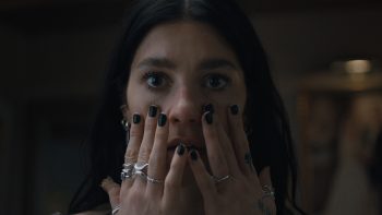 Le visage d'une femme apparaît en gros plan ; elle a les yeux écarquillés et portent les mains sur ses joues ; ses ongles sont couverts de vernis noir et elle porte plusieurs bagues aux doigts ; l'arrière-plan est flou, mais surtout plongé dans l'obscurité ; plan de la Saison 1 de UN TRES MAUVAIS PRESSENTIMENT, série Netflix.