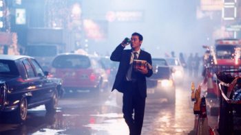 un homme élégant marche dans la rue la nuit, une biere dans la main droite, un bucket dans la main gauche, entouré de voitures et de caddies ; plan issu de running out of time.