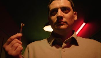 Un homme filmé en contreplongée tient une clé dans la main gauche ; il la brandit du bout des doigts ; son regard est à la fois sombre et déterminé ; dans son dos, on devine un néon rouge et un autre plafonnier ; le plafond est pour le reste entièrement dans l'obscurité ; plan du film PAST LIFE, projeté au BIFFF 2026. 