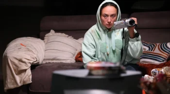 Une jeune femme assise sur un canapé porte un hoodie vert à capuche et tient dans sa main gauche une caméra DV. 