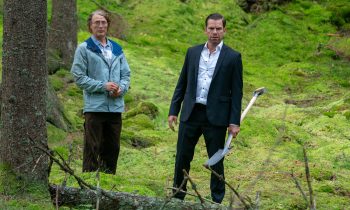 Deux hommes se tiennent debout au milieu d'une forêt, de jour ; celui de gauche porte un manteau bleu, celui de droite un costume trois pièces ; celui de droite tient une pelle dans la main gauche ; plan du film THE LAST VIKING, projeté au Grindhouse Paradise 2026.