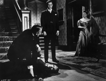 Dans le hall d’une vieille maison, au pied des escaliers, un homme inspecte le cadavre d’une femme ; celle -ci est certainement tombée des escaliers devant le regard médusé de deux personnes, une femme en arrière-plan, un homme au milieu du plan ; l'image est en noir et blanc ; plan du film LE SPECTRE DU CHAT de la Hammer.