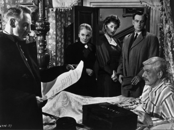 Dans la chambre d’une vieille maison victorienne, un homme lit un parchemin à un autre alité en pyjama ; dans cette pièce, on aperçoit aussi trois autres personnes, deux femmes et un homme, qui assistent à la scène ; l'image est en noir et blanc ; plan du film LE SPECTRE DU CHAT de la Hammer.