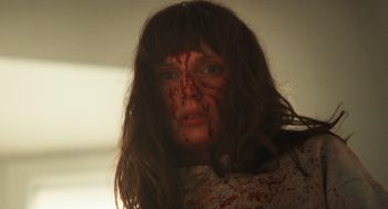 Une femme aux longs cheveux châtains regarde vers nous en contre-plongée ; elle a le visage maculé de sang ; elle affiche un air inquiet ; derrière elle, on devine des murs et un plafond blancs ; plan du film PLUS FORT QUE LE DIABLE, projeté au BIFFF 2026.