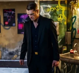 Un homme élégant, tout de noir vêtu, tient un sac en cuir dans sa main, s'apprêtant à quitter les toilettes de ce qui semble être un bar, dont les murs sont taggés ; plan issu de la série Hary Hole saison 1.