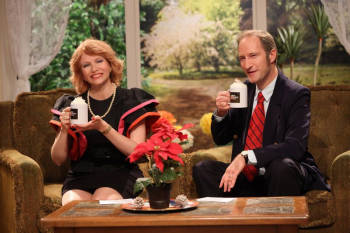 Une femme rousse avec une robe noir et rouge assise a coté d'un homme en costard cravate tiennent tous deux un mug dans un décor évoquant celui des morning show américain