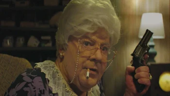 Une grand-mère au visage masculin brandit un petite revolver de type Derringer ; on suppose qu'il s'agit d'un homme déguisé en femme ; la grand-mère a une cigarette en bouche, des lunettes, des boucles d'oreille et des cheveux blancs ; son regard est inquiet ; autour d'elle, on imagine un salon, avec un petit abat-jour qui éclaire la pièce ; plan du film FUCK MY SON!, projeté au BIFFF 2026.