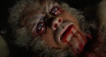 Un loup garou aux poils grisonnant agonisant au sol avec du sang qui lui sort de la bouche