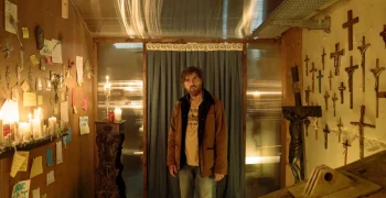 Un homme vient d'entrer dans une pièce éclairée par des bougies ; il n'y a aucune fenêtre et la lumière est juste celle qui émane des bougie ; de nombreux crucifix sont accrochés à un des murs ; il regarde vers nous, tandis qu'un rideau se trouve dans son dos, obstruant une ouverture ; plan du film THE HOLY BOY, projeté au BIFFF 2026.