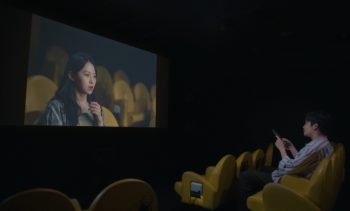 Un garçon est assis dans une salle de cinéma plongée dans l'obscurité ; les sièges sont jaunes ; il regarde l'écran sur lequel apparaît le visage d'une jeune femme ; la jeune femme sur l'écran est elle aussi dans une salle de cinéma avec des sièges jaunes ; plan du film YOU ARE THE FILM, projeté au BIFFF 2026.