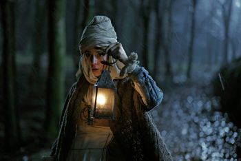 Une jeune femme au visage inquiet avance dans les bois de nuit ; elle porte une coiffe blanche faite d'un haut turban et tient une lanterne allumée dans la main gauche ; la forêt est plongée dans l'obscurité de la nuit, tandis qu'elle se tient sur un large sentier ; à ses vêtements, on comprend que la scène doit avoir lieu au 17e siècle ; plan du film GAUA, projeté au BIFFF 2026.