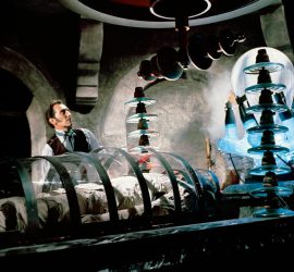 Frankenstein joué par Peter Cushing dans son laboratoire, devant sa créature encore endormie dans une boite en verre, entouré de tubes et d'objets scientifiques.
