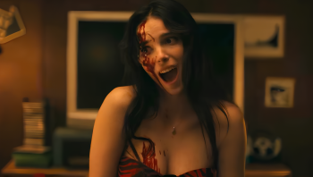 Une jeune femme aux longs cheveux noirs pousse un cri, le visage légèrement distordu ; du sang lui couvre la partie droite de la face et macule le haut de sa poitrine ; elle semble porte une robe rouge, les bras dénudés ; derrière elle, on devine le mur d'une pièce, avec des photos épinglées et peut-être une télévision ; plan du film OBSESSION de Curry Barker.