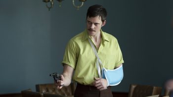 Un homme avec le bras gauche en écharpe braque un revolver face à lui ; il porte une moustache, les cheveux courts et est vêtu d'une chemise ; derrière lui, on aperçoit un mur gris monochrome et on devine des sièges en cuir ; plan du film LA CORDE AU COU de Gus Van Sant.