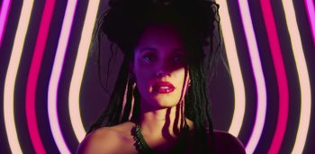 Une femme noire est filmée à hauteur d'épaules ; elle porte des cheveux noirs tressés et semble maquillée ; derrière elle, on aperçoit des sortes de néons colorés dans les tons roses et jaunes ; son visage est partiellement dans l'ombre, tandis qu'elle regarde droit devant elle ; plan du film LOVE KILLS, projeté au BIFFF 2026.