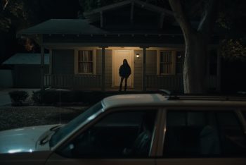 De nuit, la silhouette ombragée d'une femme apparaît au loin sous le porche d'une maison en arrière-plan ; à l'amorce du plan, on aperçoit une voiture, garé à quelque distance de la maison en bois ; un arbre apparaît sur la droite entre la voiture et l'ombre de la jeune femme ; plan du film OBSESSION de Curry Barker.