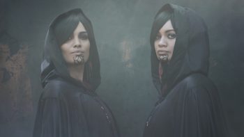 Deux femmes maories se tiennent face à face et ont la tête tournée vers la caméra ; elles portes toutes deux un vêtement noir avec un capuchon rabattu ; elles ont la même coupe de cheveux et un tatouage sur le menton ; derrière elle, la brume domine et on peine à reconnaître le moindre paysage ; plan du film TAUMANU de Taratoa Stappard.